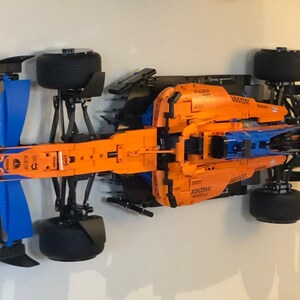 Puede incluir: Un modelo Lego detallado de un coche de carreras de F&oacute;rmula 1 McLaren. El coche es naranja y azul con detalles negros. El modelo se muestra en una pared blanca.