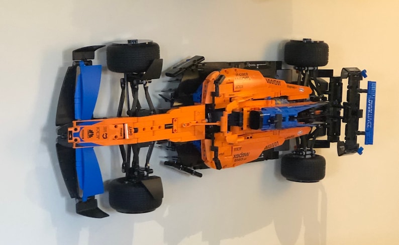 Puede incluir: Un modelo Lego detallado de un coche de carreras de F&oacute;rmula 1 McLaren. El coche es naranja y azul con detalles negros. El modelo se muestra en una pared blanca.