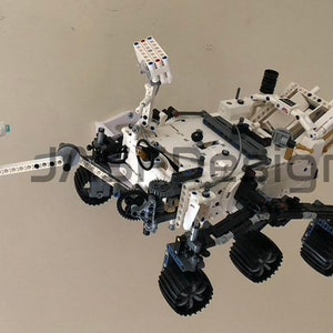 May include: A white and black Lego model of the Mars rover Curiosity. The rover has six wheels, a robotic arm, and a camera on a mast. The model is detailed and realistic.