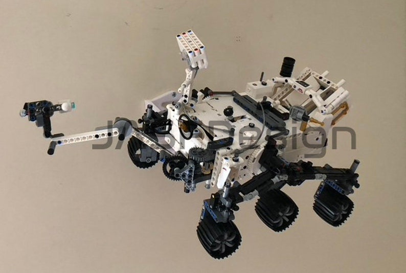 May include: A white and black Lego model of the Mars rover Curiosity. The rover has six wheels, a robotic arm, and a camera on a mast. The model is detailed and realistic.