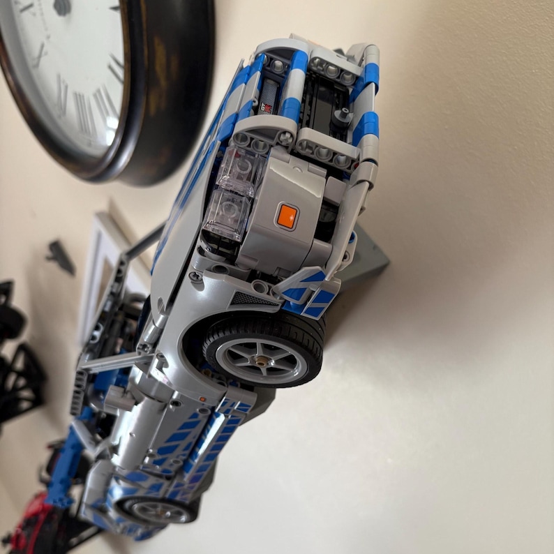 May include: A silver and blue toy car model with black wheels. The model has blue stripes and details, and is made of interlocking plastic bricks. The car is displayed on a wall, near a clock.