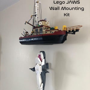 May include: A Lego Jaws wall mounting kit featuring a boat and a shark. The boat is red, beige, and black, with a tall mast. The shark is white and gray, with an open mouth. Text on the image reads "Lego Jaws Wall Mounting Kit" and "Lego not Include".