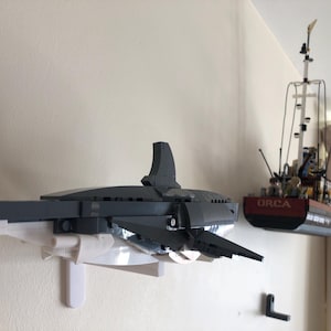 May include: A Lego model of a gray and white shark mounted on a white bracket, with a Lego boat model to the right. The boat is red and black with the word "ORCA" on the side. The models are mounted on a white wall.