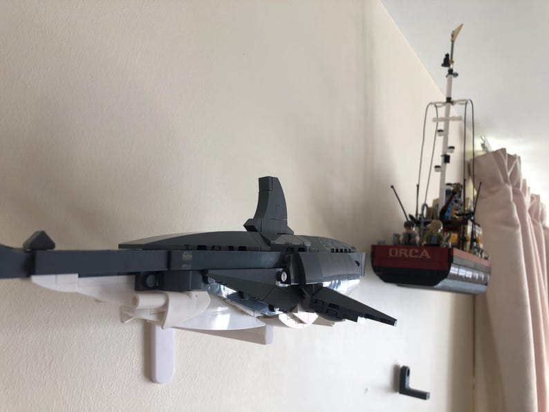 May include: A Lego model of a gray and white shark mounted on a white bracket, with a Lego boat model to the right. The boat is red and black with the word "ORCA" on the side. The models are mounted on a white wall.