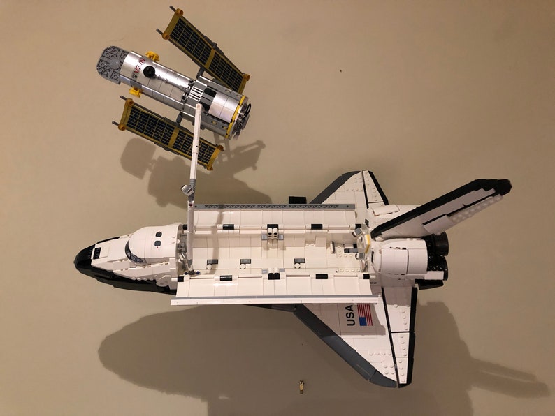 Puede incluir: Un modelo Lego blanco y negro de una lanzadera espacial con la palabra "USA" impresa en el lateral. La lanzadera lleva un modelo Lego blanco y plateado del telescopio espacial Hubble.