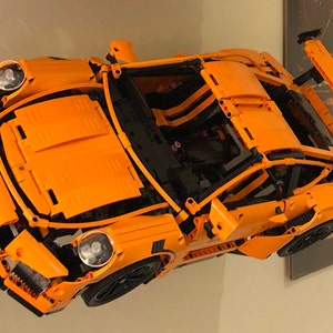 May include: An orange Lego Porsche 911 GT3 RS model car with black accents and a rear spoiler. The car is built with Lego bricks and is displayed on a wall.