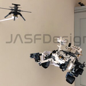 May include: A white and gray Lego model of the Perseverance rover, a Mars exploration vehicle, is suspended from the ceiling. The rover has a robotic arm, wheels, and a camera. The model is suspended from the ceiling by a white arm attached to a black and gray Lego helicopter.