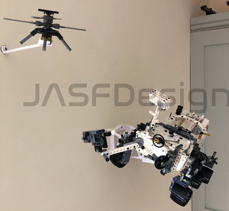May include: A white and gray Lego model of the Perseverance rover, a Mars exploration vehicle, is suspended from the ceiling. The rover has a robotic arm, wheels, and a camera. The model is suspended from the ceiling by a white arm attached to a black and gray Lego helicopter.