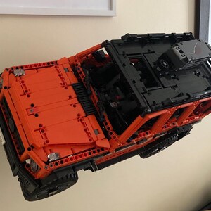 May include: A bright orange and black Lego model of a Mercedes-Benz G-Wagon SUV. The model is suspended in mid-air, as if it is floating.
