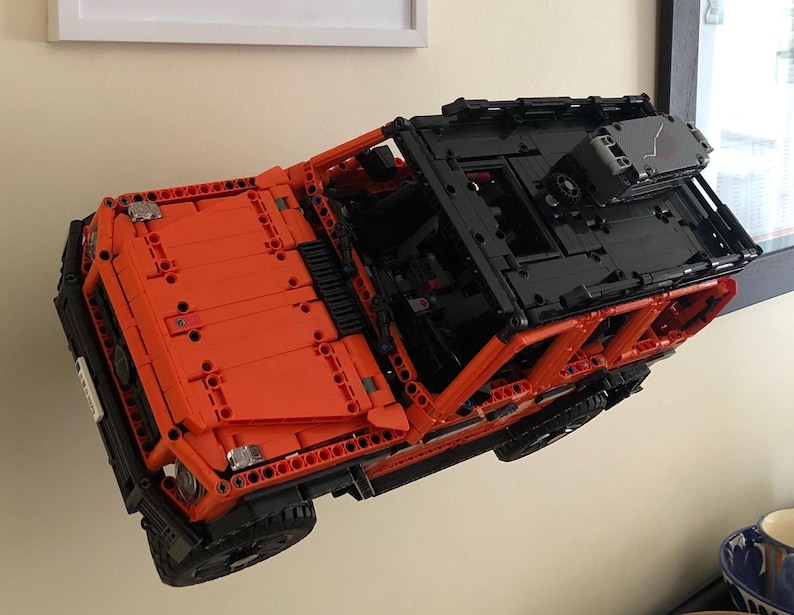 May include: A bright orange and black Lego model of a Mercedes-Benz G-Wagon SUV. The model is suspended in mid-air, as if it is floating.