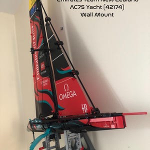 K&ouml;nnte beinhalten: Ein rotes, schwarzes und wei&szlig;es Modell der Emirates Team New Zealand AC75 Yacht (42174) ist an einer grauen Wandhalterung montiert. Das Modell besteht aus LEGO-Steinen und enth&auml;lt das Logo des Teams sowie Sponsoren. Der Text "Lego Not Included" ist unter dem Modell aufgedruckt.