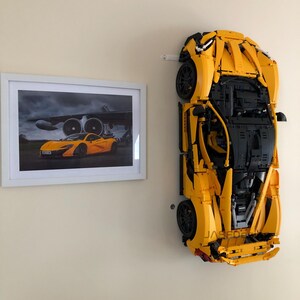 May include: A large, yellow Lego model of a sports car mounted on a wall, next to a framed picture of a similar car. The Lego car is detailed with black accents and wheels. The picture shows the car in front of an airplane.