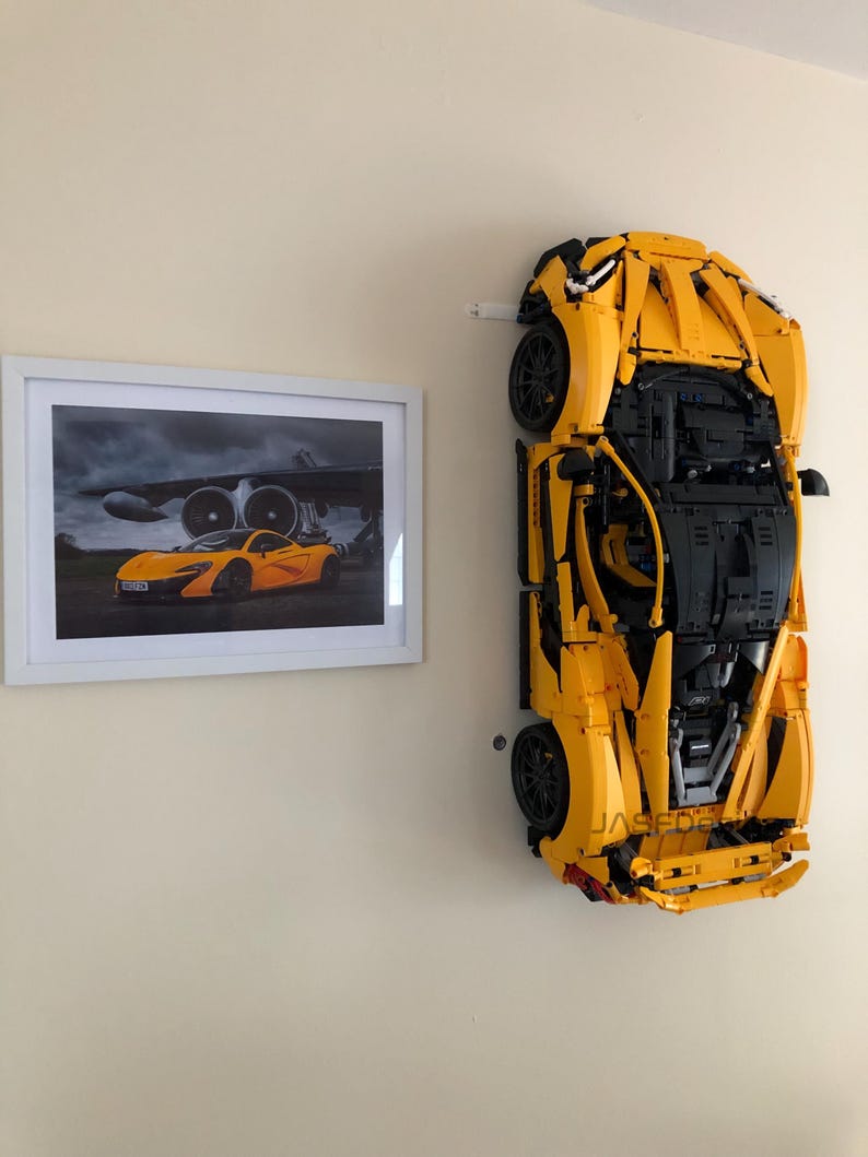 May include: A large, yellow Lego model of a sports car mounted on a wall, next to a framed picture of a similar car. The Lego car is detailed with black accents and wheels. The picture shows the car in front of an airplane.