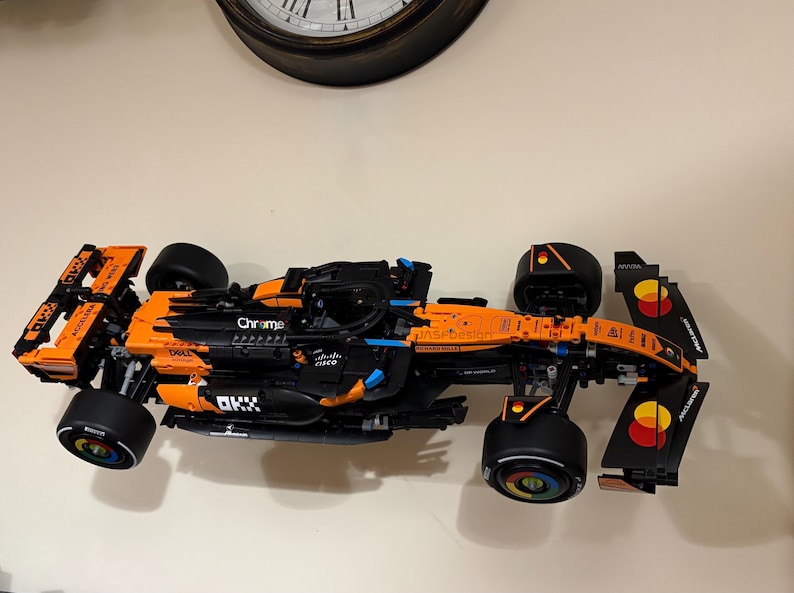 May include: A black and orange race car model with sponsor logos, including "Chrome" and "Dell." The model has large black tires and is set against a neutral background. The car's design is detailed, with visible mechanical components.