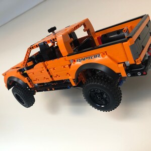 Op de afbeelding: Een oranje Lego Ford Raptor pick-up truck met zwarte wielen en het woord "RAPTOR" erop gedrukt. De truck hangt in de lucht tegen een witte muur.
