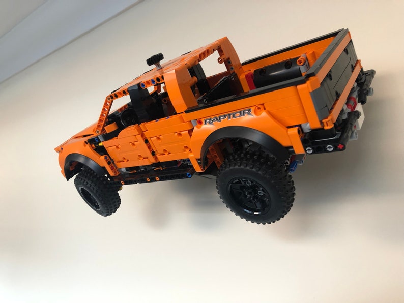 Op de afbeelding: Een oranje Lego Ford Raptor pick-up truck met zwarte wielen en het woord "RAPTOR" erop gedrukt. De truck hangt in de lucht tegen een witte muur.