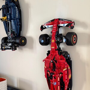 May include: Two Lego Formula 1 race cars mounted on a wall. The red car is a Ferrari with the Shell logo and the blue car is a Red Bull with the Oracle logo.