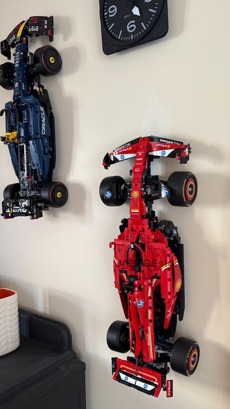 May include: Two Lego Formula 1 race cars mounted on a wall. The red car is a Ferrari with the Shell logo and the blue car is a Red Bull with the Oracle logo.
