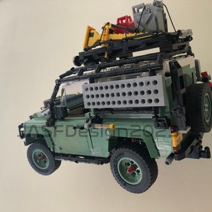 May include: A green Lego Land Rover Defender with a black roof rack and a variety of accessories, including a yellow shovel, a red gas can, and a gray water container. The vehicle is suspended in mid-air, as if it is floating.