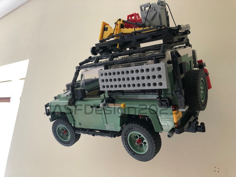 May include: A green Lego Land Rover Defender with a black roof rack and a variety of accessories, including a yellow shovel, a red gas can, and a gray water container. The vehicle is suspended in mid-air, as if it is floating.