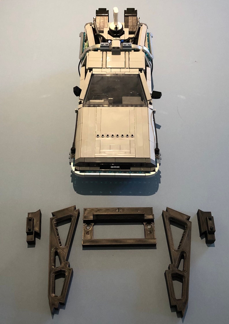 May include: A white Lego DeLorean time machine from the Back to the Future movie franchise. The car is on a blue surface with four gray Lego pieces beside it.