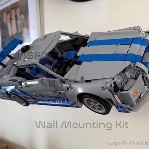 May include: A gray and blue model car made of interlocking plastic bricks, mounted on a wall. The car has blue stripes and accents. The text "Wall Mounting Kit" is visible below the car.