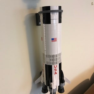 May include: A white and black LEGO model of the Saturn V rocket with the words "USA" printed on the side. The rocket is mounted on a wall with a black and gray bracket.