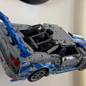 May include: A gray and blue model car with a detailed design. The car features blue accents, including a spoiler and side detailing. The wheels are visible, and the car appears to be a replica of a sports car.
