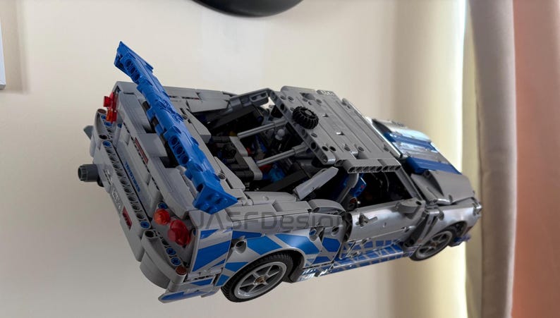May include: A gray and blue model car with a detailed design. The car features blue accents, including a spoiler and side detailing. The wheels are visible, and the car appears to be a replica of a sports car.