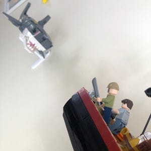 May include: A Lego scene depicting a shark attacking a ship. The shark is gray and white with a wide open mouth. Two Lego figures are on the ship, one holding a sword. The ship is red, black, and brown.