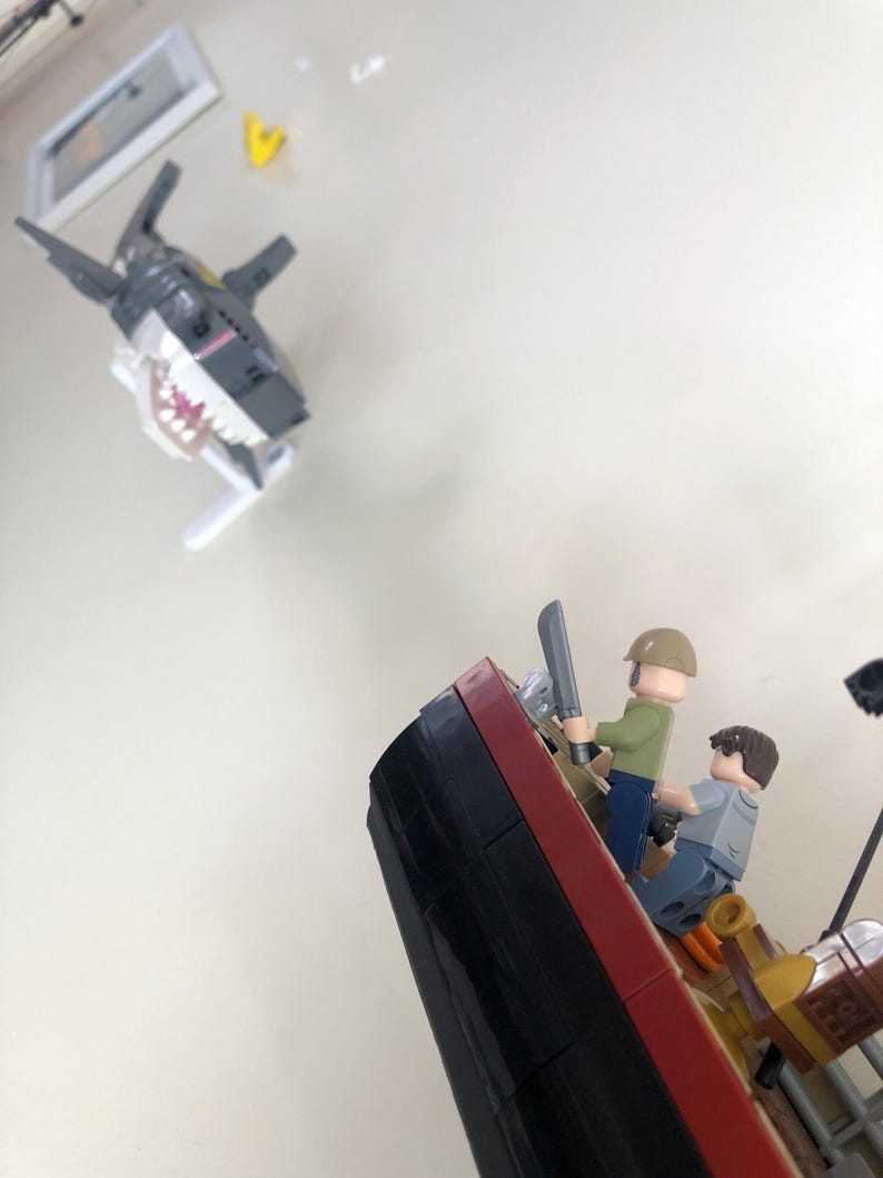 May include: A Lego scene depicting a shark attacking a ship. The shark is gray and white with a wide open mouth. Two Lego figures are on the ship, one holding a sword. The ship is red, black, and brown.