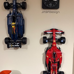 May include: Two Lego Formula 1 race cars mounted on a wall. The car on the left is blue and black with red accents and the car on the right is red with black and white accents. The cars are displayed upside down with the wheels facing the wall.