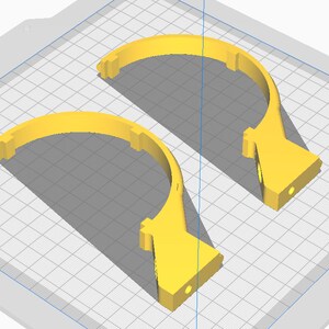 May include: Two yellow, semi-circular 3D-printed objects. Each piece has a flat base with a circular hole and a curved top with rectangular notches. The objects are set against a grid background, suggesting a design or modeling context.