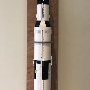 May include: A white and black Lego model of the Saturn V rocket, mounted on a wooden board. The rocket has the words "United States" printed on the side, and the American flag is visible on the side of the rocket.
