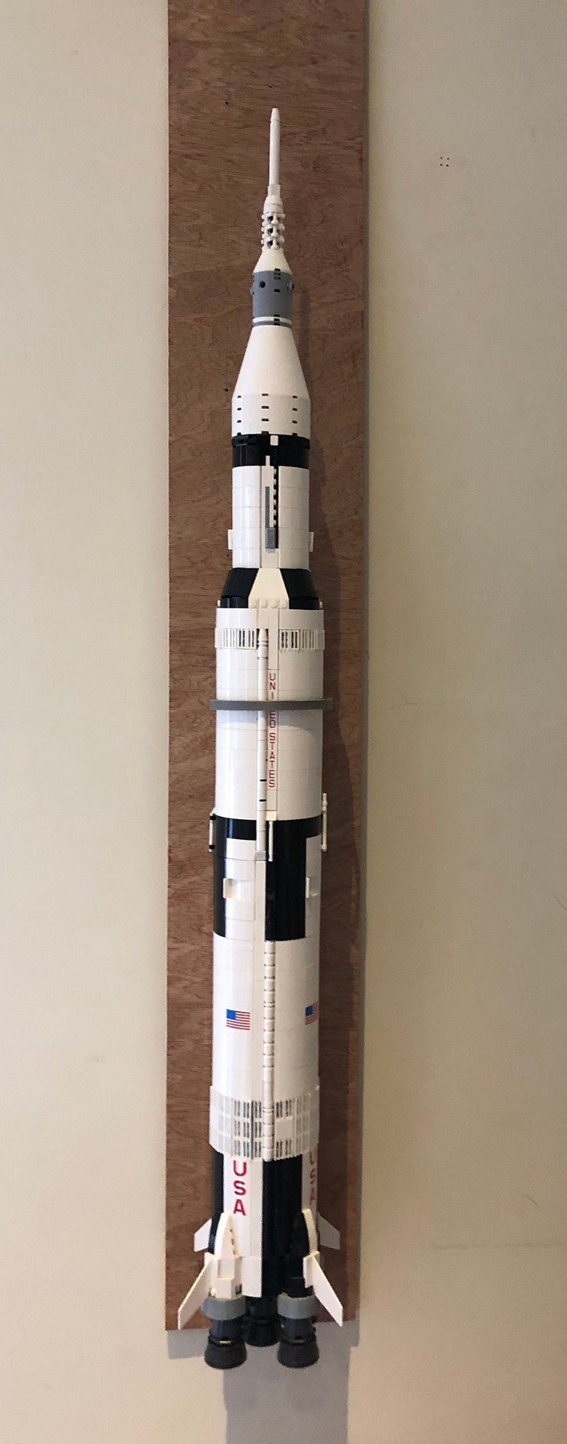 May include: A white and black Lego model of the Saturn V rocket, mounted on a wooden board. The rocket has the words "United States" printed on the side, and the American flag is visible on the side of the rocket.