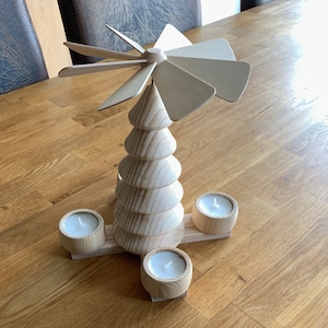 Può includere: Una piramide di Natale in legno con un'elica bianca e tre portacandele. La piramide è realizzata in legno naturale e ha un aspetto rustico.