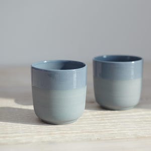 Puede incluir: Dos tazas de cerámica con un esmalte azul claro en la mitad superior y un esmalte blanco en la mitad inferior. Las tazas están sobre una superficie de madera clara.