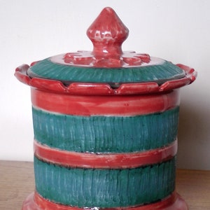 May include: A red and green ceramic jar with a lid. The jar has a scalloped rim and a decorative design. The lid has a knob on top.