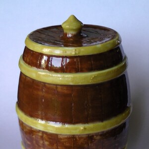 May include: A ceramic barrel-shaped jar with a lid. The jar is painted to look like wood with brown and yellow stripes.