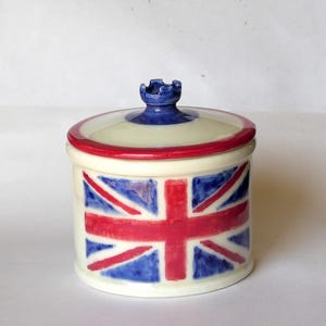 May include: A cylindrical ceramic canister with a cream-coloured base featuring a hand-painted Union Jack flag design in red and blue. The lid has a matching red rim and a small, dark blue knob handle at the centre with a crown-like shape.