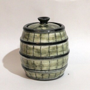 May include: A ceramic barrel-shaped container with a lid. The container is painted in shades of green and grey, with black bands and vertical lines to mimic a wooden barrel. The lid has a small, dark knob.