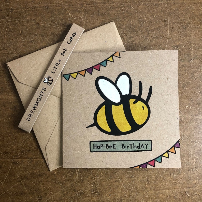 May include: A brown card with a yellow and black bee illustration and the text "Hap-Bee Birthday". The bee is flying with a string of colorful flags above it.