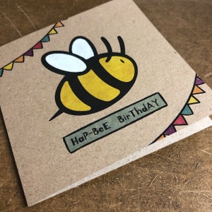 May include: A brown card with a yellow and black bee illustration and the text "Hap-Bee BirThday" in a green banner. The card has a colorful string of pennants at the top.