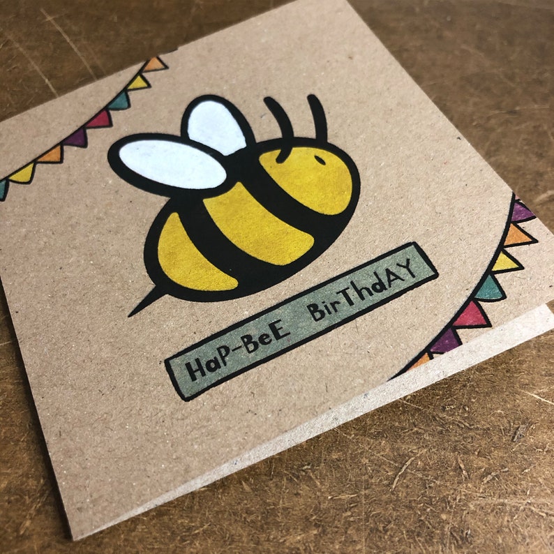 May include: A brown card with a yellow and black bee illustration and the text "Hap-Bee BirThday" in a green banner. The card has a colorful string of pennants at the top.