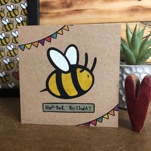 May include: A brown card with a cartoon bee and colorful bunting. The card says "Hap-Bee Birthday" in a green banner.