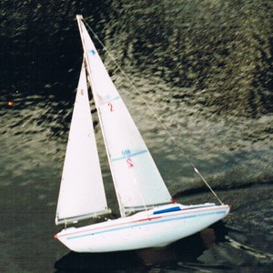 May include: A white sailboat with a red and blue stripe on the hull, sailing on a dark body of water. The sail is white with a blue stripe and the number "2" printed on it.