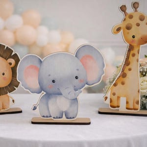 Puede incluir: Tres recortes de animales de madera: un león con melena marrón, un elefante gris con orejas rosas y una jirafa con manchas marrones. Están colocados sobre un mantel blanco, con globos y flores en el fondo. Ideal para un baby shower.