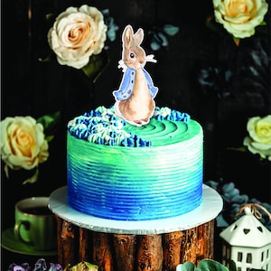 May include: A blue and green ombre cake with a Peter Rabbit cake topper. The cake is on a wooden cake stand.