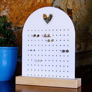 May include: White earring holder with a heart cutout and a wooden base. The holder has many small holes to hold earrings.