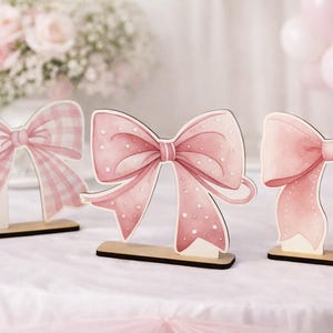 Könnte beinhalten: Drei rosa Schleifendekorationen auf Holzsockeln. Die Schleifen haben verschiedene Designs, darunter ein Vichy-Muster und Tupfen. Der Hintergrund umfasst Blumen, Kerzen und rosa Luftballons, wodurch eine weiche, feminine Ästhetik entsteht.